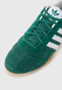 Sapatilha atlética verde com textura acolchoada, detalhes brancos em três riscas e sola de borracha. Materiais em camurça e sintético são utilizados.