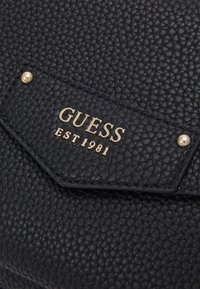 Sort tekstureret læderhåndtaske med et centralt skilt med "GUESS" og "EST 1981" i guld, accentueret af to guldnitter.