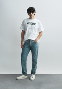 Weißes Baumwoll-T-Shirt mit schwarzem "MOSCHINO Milano"-Schriftzug, kombiniert mit hellblauen Jeans und weißen Sneakers. Accessoires sind schwarze Sonnenbrillen.