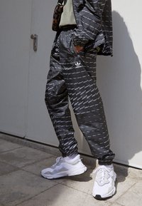 Svarta träningsbyxor med vit upprepande "adidas"-logotyp, elastiska muddar och sidofickor, som matchas med vita nätadidasskor.