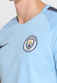 Helles blaues Nike-T-Shirt mit V-Ausschnitt und einem Manchester City-Logo auf der linken Brust. Verfügt über schwarze Akzente an den Schultern. Weiche Textur.