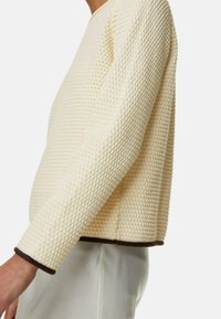 Teksturowany sweter w kolorze kremowym o luźnym kroju, z plecionym wzorem i ciemnobrązowymi wykończeniami przy mankietach i u dołu. Miękki materiał.