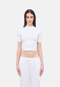 Weißes, cropped Oberteil mit kurzen Ärmeln und hohem Kragen, das eine glatte Textur aufweist. Kombiniert mit weiten weißen Jogginghosen mit Kordelzug und Taschen.