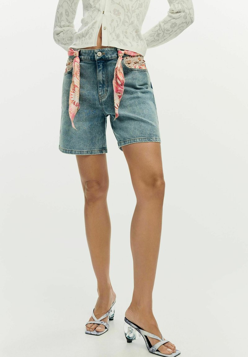 Femme portant un short en jean avec des bandanas floraux noués sur les poches, un cardigan court blanc à motifs et des sandales à talons argentées.
