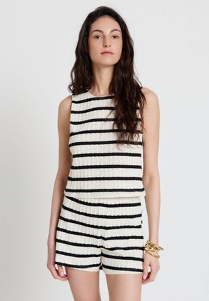 Femme aux longs cheveux bruns portant un débardeur en maille et un short à rayures horizontales noires et blanches, debout devant un fond blanc uni.