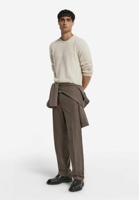 Jasno beżowy sweter dzianinowy, brązowe dopasowane spodnie i czarne buty. Sweter zarzucony na ramionach, tworząc warstwowy wygląd. Gładkie faktury.