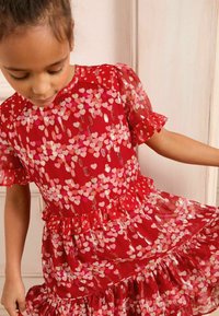 Rotes Kleid mit kurzen, gepufften Ärmeln, versehen mit gestaffelten Rüschen und einem herzförmigen Design in Pink und metallischen Akzenten. Transparenter Stoff.