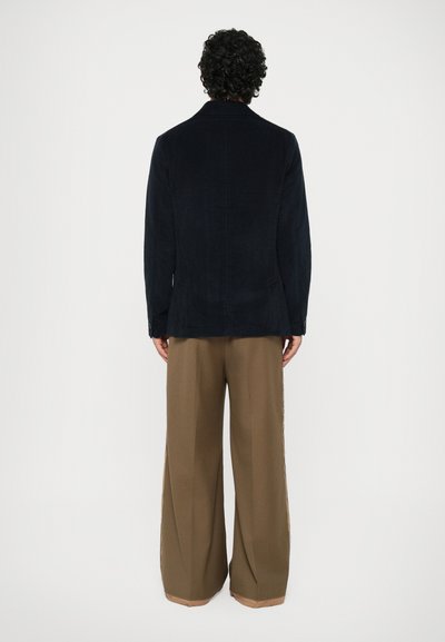 Sacou din catifea de culoare bleumarin, cu croială dreaptă, rever în v, și două butoane, purtat cu pantaloni largi maro, cu tiv bej.