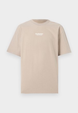 Beež puuvillane T-särk lühikeste varrukatega ja ümmarguse kaelusega. Ees on valge "Hollister Original Designs" logo.