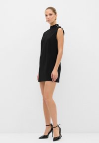 Vestido negro sin mangas con cuello alto simulado, botones laterales y un corte recto, combinado con tacones negros de punta y correas en el tobillo.