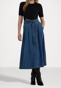 Denimová maxi sukně s pásem na zavazování, s plisovaným designem a dvěma bočními kapsami. V kombinaci s černým texturovaným topem s krátkými rukávy a kozačkami do kolen.