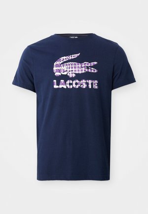 T-shirt in cotone blu navy con maniche corte, caratterizzato da un logo a coccodrillo rosa e bianco e la scritta "LACOSTE" sotto.