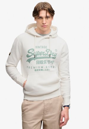 Ung mand iført en off-white SuperDry hættetrøje med frontlomme og beige bukser, poserer med den ene hånd i lommen mod en ensfarvet baggrund.