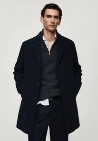 Cappotto di lana blu navy con colletto a revere, design monopetto e due tasche frontali, indossato sopra un maglione scuro e una camicia a righe.