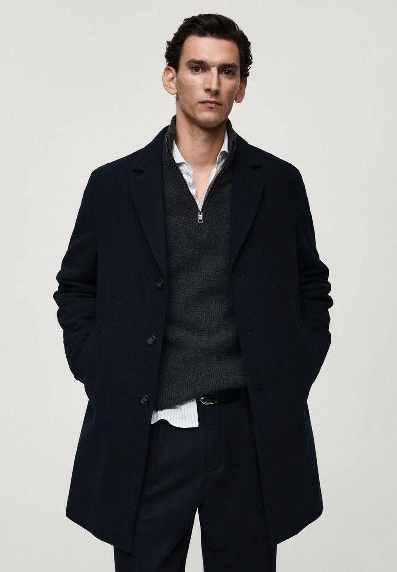 Cappotto di lana blu navy con colletto a revere, design monopetto e due tasche frontali, indossato sopra un maglione scuro e una camicia a righe.