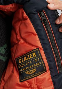 Navy- und orangefarbene gesteppte Jackeninnenseite mit sichtbaren Nähten, schwarzem Reißverschluss und einem schwarz-goldenen Etikett mit der Aufschrift 'GLAZER' und 'PME LEGEND.'