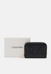 Black leather wallet with embossed "CK" logo, gold zipper detail, and a smooth texture, placed beside a white cardboard box labeled "Calvin Klein."