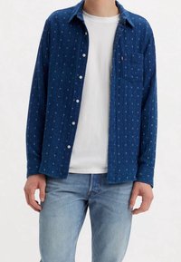 Camicia azzurra a maniche lunghe con piccoli motivi geometrici, con bottoni frontali, taschino sul petto, indossata sopra una maglietta bianca e abbinata a jeans chiari.
