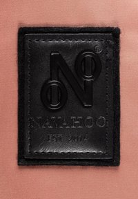 Etichetta in pelle nera goffrata con un logo circolare, "NAVAHOO," e "EST 2014," posizionata su uno sfondo in tessuto rosa testurizzato.