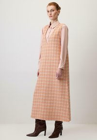 Maxi dress in orange plaid fabric with a V-neckline; layered over a light pink shirt with long sleeves. Features brown pointed ankle boots.