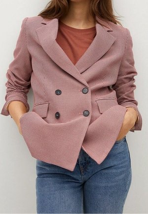 Vrouw met een dubbelgeknoopt roze-wit geruit colbert over een roestkleurige top en blauwe spijkerbroek, handen in de zakken.