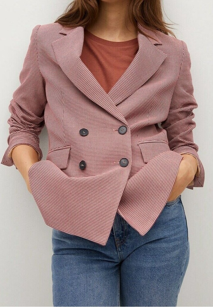 Femme portant un blazer croisé à carreaux roses et blancs sur un haut couleur rouille et un jean bleu, les mains dans les poches.