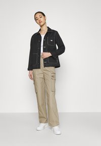 Zwarte denimjas met witte stiksels, voorzien van meerdere zakken en een logo. Gecombineerd met beige cargobroek en witte sneakers.