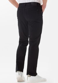 Black cotton pants with a slim fit, featuring back pockets, a smooth texture, and a subtle brand logo on the waistband, paired with white sneakers.