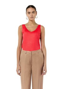 Czerwona bezrękawnik o dekolcie w kształcie litery V, zestawiony z jasnobrązowymi spodniami o wysokim stanie. Tkanina wydaje się gładka, o minimalnej teksturze.