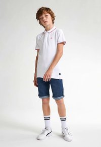 Valkoinen poolopaita punaisilla yksityiskohdilla, yhdistetty farkkushortseihin. Valkoiset tennarit ja polven korkeuteen ulottuvat sukat, joissa mustat raidat, täydentävät asun.