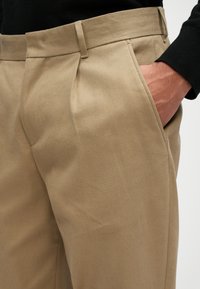 Smėliniai pritaikyti kelnių modeliai su lygiu paviršiumi, turintys dvi priekinės raukšlės, šonines kišenes ir liekną siluetą.