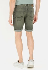 Olijfgroene denim shorts voor heren met opgerolde omslagen, voorzien van twee achterzakken en een tan leren patch op de tailleband. Witte sneakers gedragen.