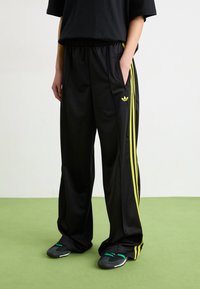 Black track pants with yellow stripes, elastic waistband, side pockets, and Adidas logo. Material appears smooth and lightweight.