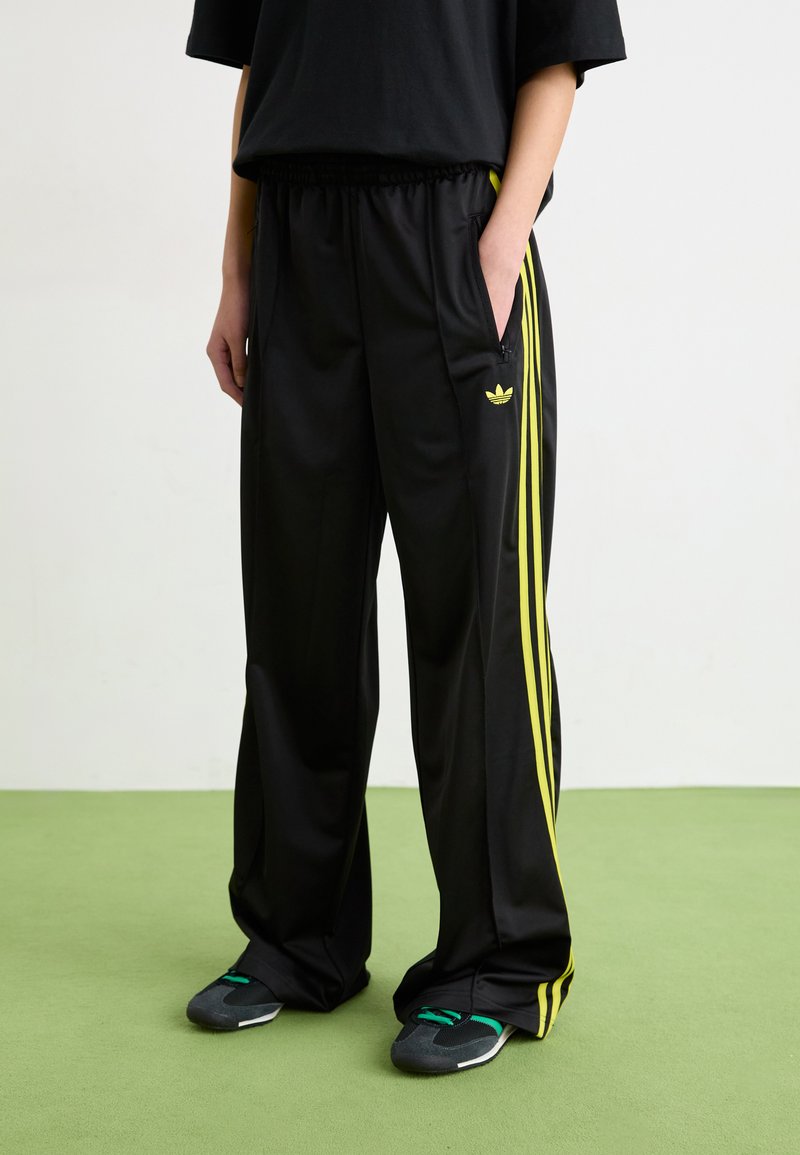 Black track pants with yellow stripes, elastic waistband, side pockets, and Adidas logo. Material appears smooth and lightweight.