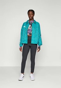 Teal mönstrad jacka med två framfickor, kombinerad med en flerfärgad kort topp och svarta leggings, avslutad med vita sneakers.