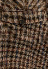 Brown and beige houndstooth fabric with a pocket, featuring small black button detail and subtle blue and burgundy lines throughout.