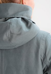 Grå hættejakke med blødt, tekstureret stof. Har en høj krave, rene linjer og detaljerede syninger langs sømmene.