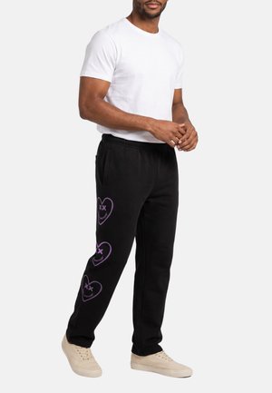 Un hombre posa con una camiseta blanca, pantalones deportivos negros con motivos de corazones morados y zapatillas beige sobre un fondo liso.