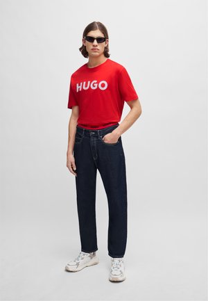 Tricou roșu din bumbac cu logo-ul alb "HUGO", blugi din denim albastru închis și adidași deschiși. Accesoriile includ ochelari de soare negri.
