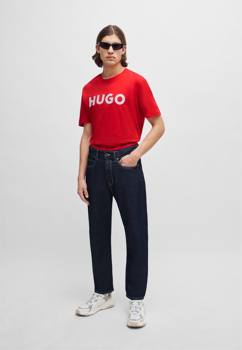 Camiseta de algodón roja con logo blanco "HUGO", jeans de mezclilla azul oscuro y zapatillas claras. Los accesorios incluyen gafas de sol negras.