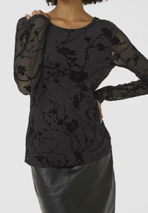 Femme portant un top noir transparent à manches longues avec des motifs floraux en velours et une jupe en cuir noire, posant avec une main sur la hanche.