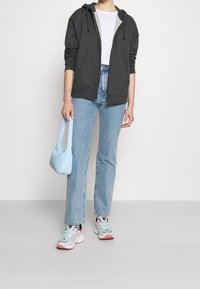 Grijze zip-up hoodie met een lichtgrijze binnenkant, gecombineerd met lichtblauwe high-waisted jeans en een pastelblauwe handtas. Witte sneakers met accenten.