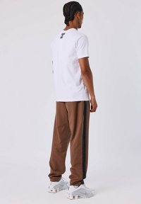 Witte t-shirt met korte mouwen en een achterkant print, gecombineerd met bruine tapse broek met een zijstrak zwarte streep, afgemaakt met witte sportschoenen.