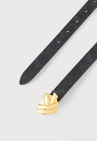 Cinturón de cuero negro con una hebilla dorada esculpida en forma de nudo sobre un fondo blanco, mostrando el extremo puntiagudo de la correa y los orificios de ajuste.