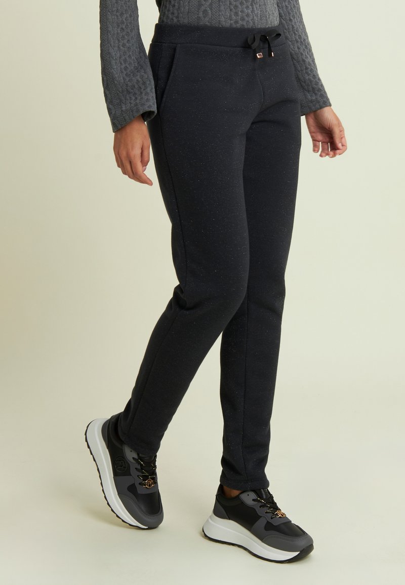 Persona che indossa pantaloni da jogging neri con macchie e sneakers grigio-neri con suola bianca, in piedi su uno sfondo chiaro.