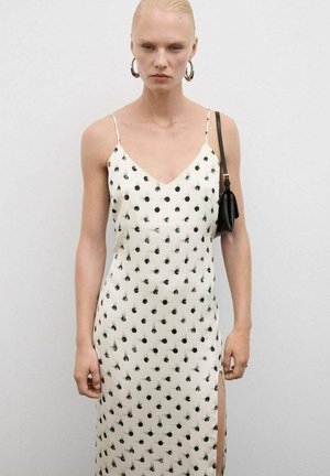 Bílé šaty s černým tečkovaným vzorem, s výstřihem do V, tenkými ramínky a postranním rozparkem, vyrobené z lehkého materiálu. Modelka drží malou černou kabelku.