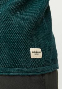 Mörk teal, stickad tröja med en strukturerad yta, ribbad nederkant och en vävd etikett med "JACK & JONES EST. 1990" på sidan.