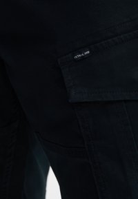 Pantaloni cargo blu scuro in tessuto robusto, dotati di multiple tasche e di un'etichetta cucita con la scritta "PETROL IND.". Texture liscia.