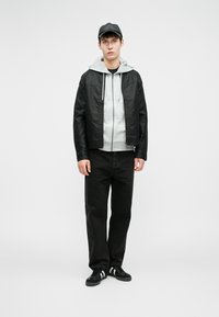 Giacca in finta pelle nera, felpa con cappuccio grigia, jeans neri e sneakers nere con strisce bianche. Il modello indossa un cappellino nero, in piedi su uno sfondo semplice.