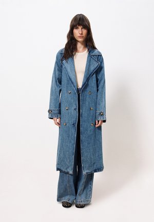 Lange denim trenchcoat met dubbelzijdige knoopsluiting, brede revers en rafelige zoom. Draagt over een beige top en flare jeans.